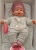 Lalka Hiszpańska 27 cm Antonio Juan 11115 Kika, bobas miękki brzuszek, płacze, mówi mama papa, bez włosów, w ubranku, magnetyczny smoczek, kołderka i poduszka